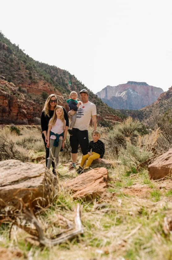 A family of five, with three children, stands outdoors on a grassy, rocky area surrounded by mountains and red rock cliffs under a bright sky. The parents hold the youngest child, and everyone faces the camera. by Justin Salem Meyer