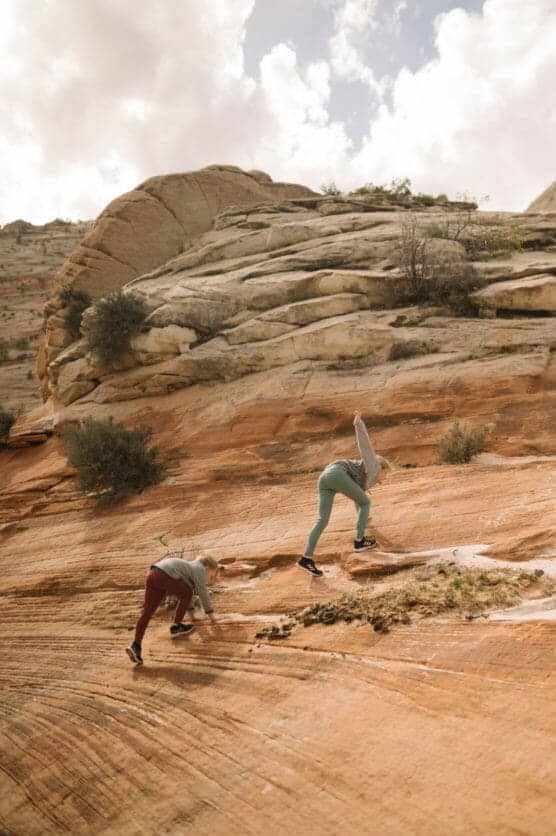 Two people wearing casual outdoor clothes are climbing up a steep, rocky sandstone slope under a partly cloudy sky, surrounded by rugged desert landscape and sparse vegetation. by Justin Salem Meyer