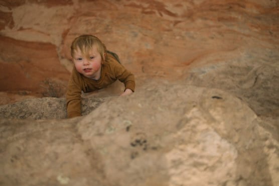 A young child with blond hair wearing a brown long-sleeve shirt climbs a large, rough rock outdoors, with reddish rock formations in the background. The child looks up while gripping the rock. by Justin Salem Meyer