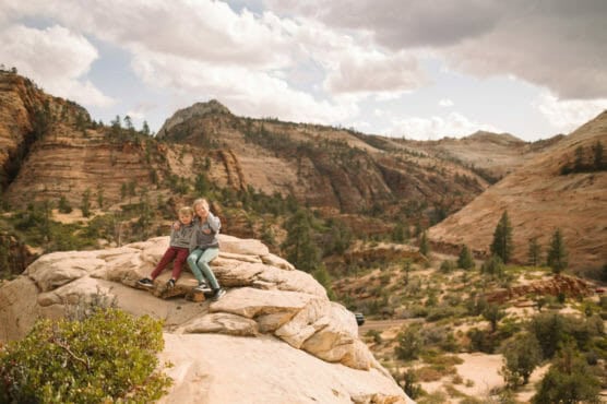 Two young children sit on a rocky outcrop overlooking a vast, rugged desert landscape with layered red rock formations, scattered trees, and a cloudy sky in the background. by Justin Salem Meyer