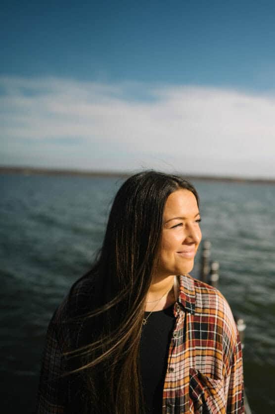 A woman with long brown hair wearing a plaid shirt stands near a body of water, smiling and looking to the right under a clear blue sky. by Justin Salem Meyer
