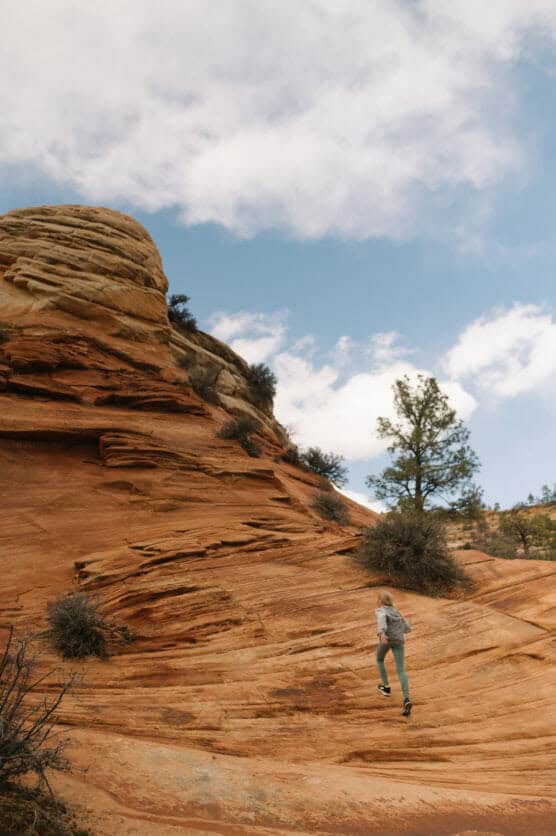 A person in outdoor clothing climbs up a sloped, orange sandstone formation under a partly cloudy sky, with small shrubs and a lone pine tree nearby. by Justin Salem Meyer