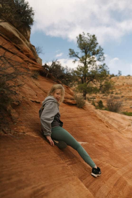 A young person in a gray hoodie and green leggings sits on a sloped, red rock surface outdoors, surrounded by desert vegetation and under a partly cloudy sky. by Justin Salem Meyer