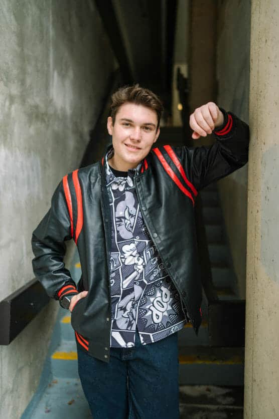 A young man with short brown hair leans against a concrete wall indoors, smiling. He wears a black leather jacket with red stripes over a patterned button-up shirt and dark blue jeans. Stairs are visible behind him. by Justin Salem Meyer
