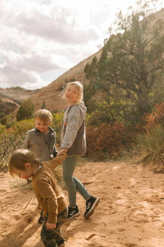 Three young children play together on a sandy trail surrounded by desert vegetation and hills under a partly cloudy sky, with one girl smiling at the camera while holding hands with the others. by Justin Salem Meyer
