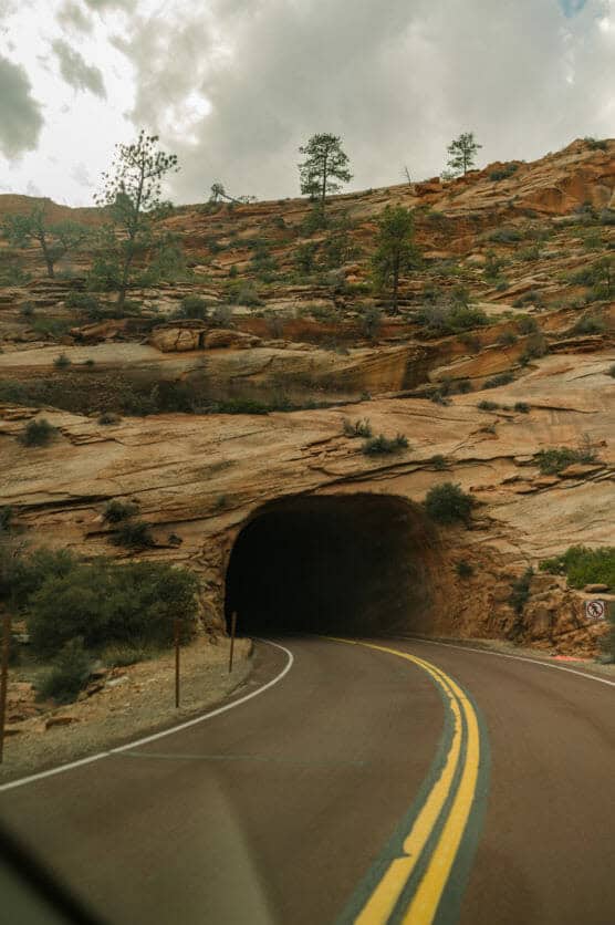 A winding road with double yellow lines leads into a tunnel carved through a reddish rocky hillside, surrounded by scattered shrubs and trees under a cloudy sky. by Justin Salem Meyer