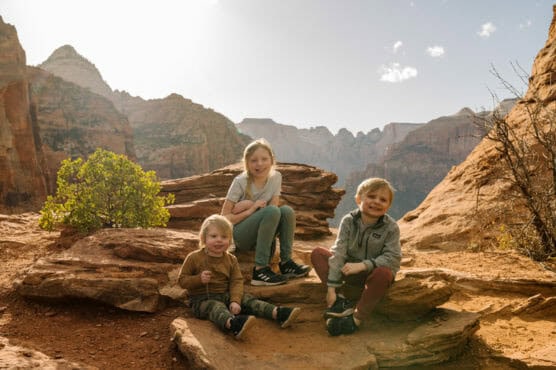 Three young children sit on large rocks in a scenic desert canyon landscape, smiling at the camera. Sunlight illuminates the rocky terrain and distant mountains in the background. by Justin Salem Meyer