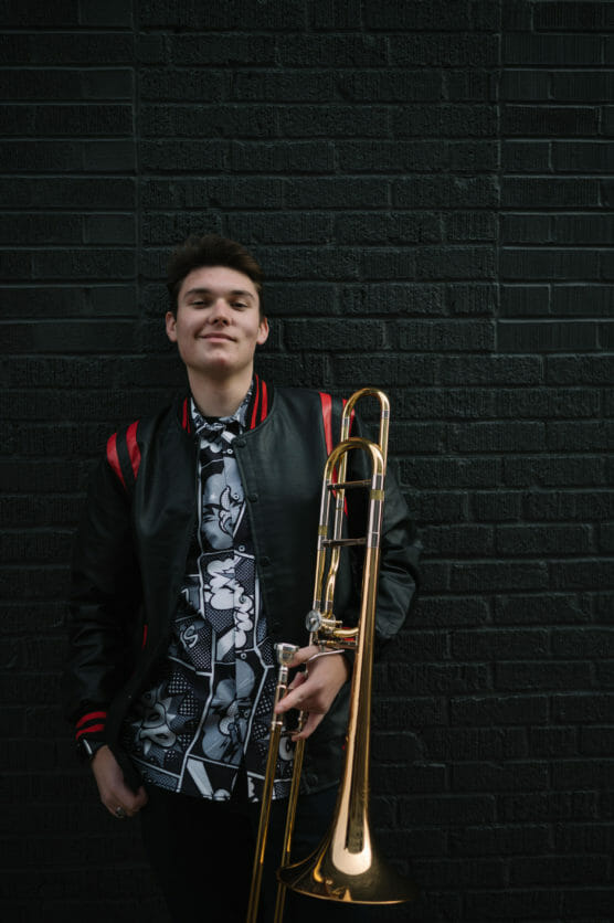 A young man in a black and red jacket stands against a dark brick wall, holding a brass trombone and smiling slightly. He wears a patterned black-and-white shirt underneath his jacket. by Justin Salem Meyer