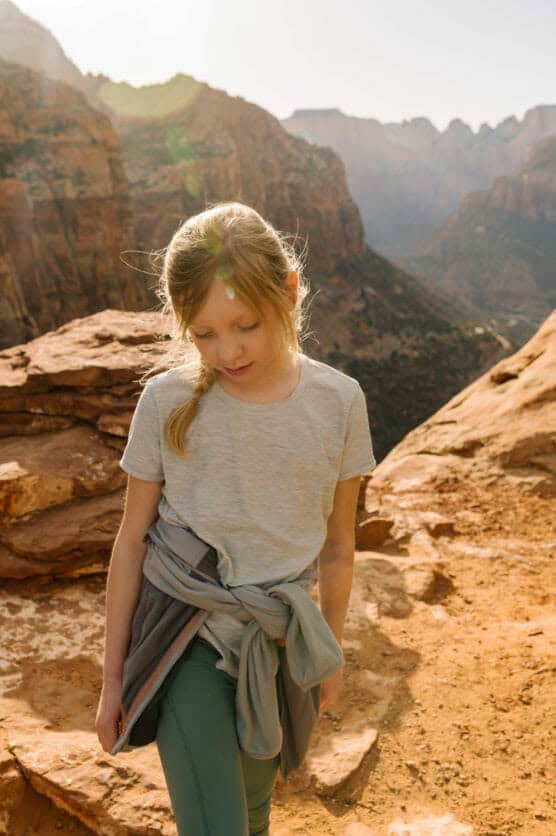 A young girl with blonde hair in a ponytail stands on a rocky cliff in a canyon, wearing a gray T-shirt and sweater tied around her waist. Sunlight shines in the background, highlighting the rugged landscape. by Justin Salem Meyer