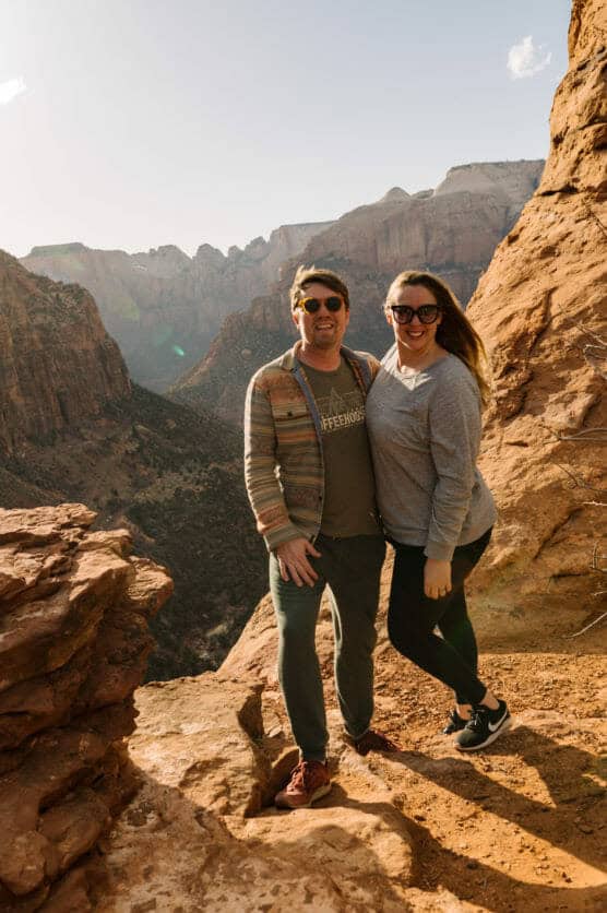 A man and woman pose and smile on a rocky cliff overlooking a scenic canyon with rugged cliffs and sunlit mountains in the background. Both are dressed casually for hiking. by Justin Salem Meyer