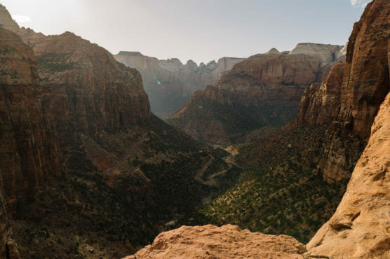 A scenic view of a wide canyon with rugged, reddish cliffs and rocky mountains under a clear sky. A winding road is visible far below, surrounded by scattered greenery. by Justin Salem Meyer