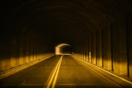 A dimly lit, empty road tunnel with two yellow lines in the center, curving gently to the right and leading toward a brighter exit in the distance. by Justin Salem Meyer