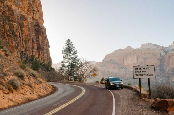 A car is parked on the side of a winding road near red rock cliffs and mountains. A road sign reads, Tunnel Ahead Watch for Stopped Vehicles. The scene is set during daylight with clear skies. by Justin Salem Meyer