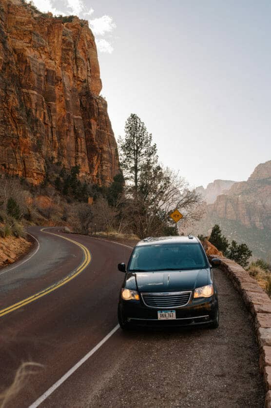 A black minivan is parked on the roadside next to a winding mountain road with tall red rock cliffs, trees, and a curve warning sign visible in the background. by Justin Salem Meyer