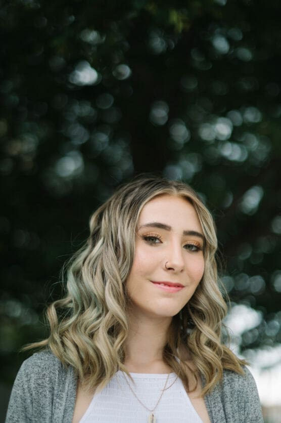 A young woman with wavy, blonde hair and a nose ring smiles softly at the camera. She is wearing a white top and gray cardigan, standing outdoors in front of a blurred, leafy background. by Justin Salem Meyer