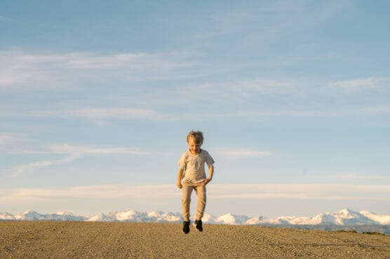 A young child is jumping in the air on a dirt path with snowy mountains in the background under a blue sky with wispy clouds. by Justin Salem Meyer