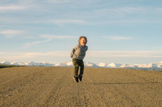 A young child in a grey sweatshirt and camouflage pants joyfully jumps on a dirt road, with snow-capped mountains and a blue sky in the background. by Justin Salem Meyer