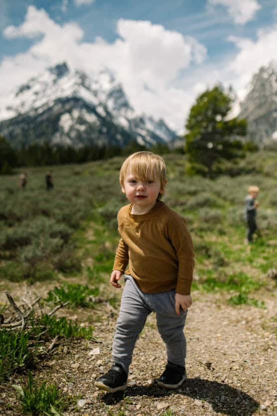 A young child walks on a gravel path in a grassy area, wearing a brown shirt and gray pants, with mountains and a partly cloudy sky in the background. Other people are blurred in the distance. by Justin Salem Meyer
