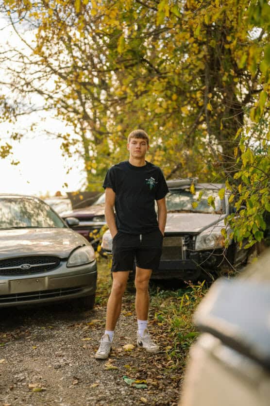 A young man wearing a black t-shirt, black shorts, and white sneakers stands on a gravel path between parked, weathered cars, surrounded by trees with green and yellow leaves. by Justin Salem Meyer