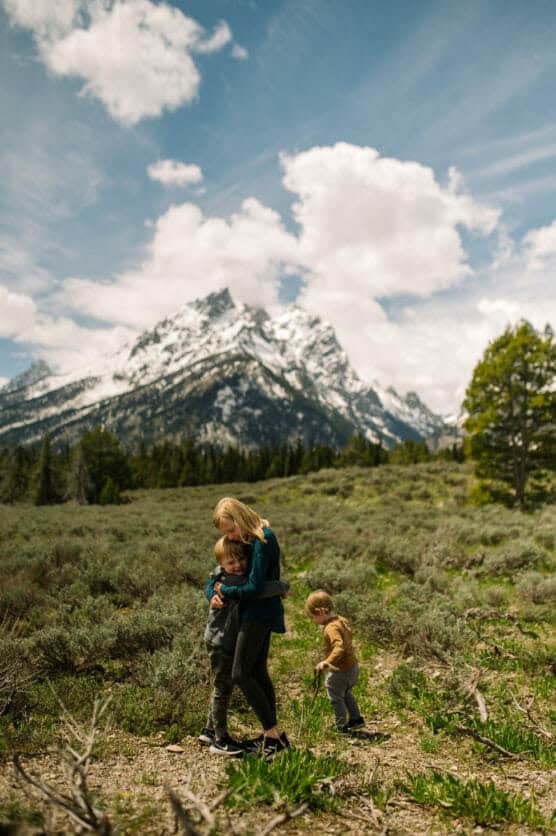 A woman hugs a young child while another child walks nearby in a grassy field with tall mountains and a partly cloudy sky in the background. by Justin Salem Meyer