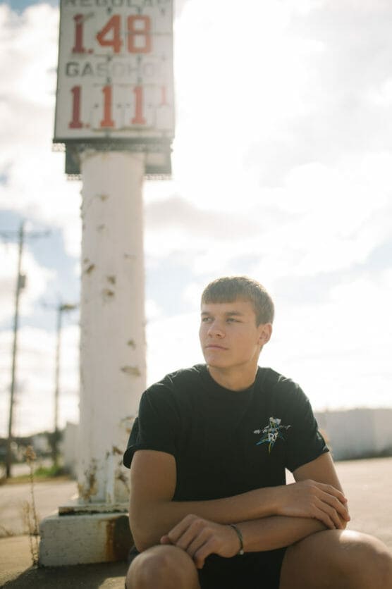 A young man in a black t-shirt sits outdoors in front of a faded, tall gas station sign showing fuel prices. The sun is bright, creating a soft, hazy background with blurred clouds and a white sky. by Justin Salem Meyer