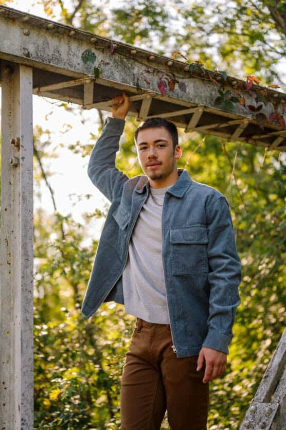 A young man wearing a blue jacket and brown pants stands outdoors, holding onto a wooden structure overhead. Green foliage and sunlight fill the background. by Justin Salem Meyer