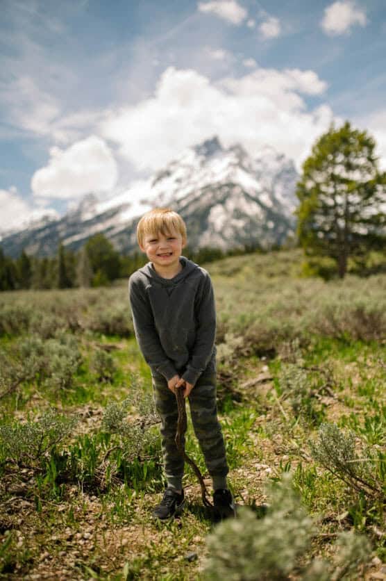 A young boy in a gray hoodie and camouflage pants stands smiling in a grassy field, holding a stick. Snow-capped mountains, trees, and a partly cloudy sky are in the background. by Justin Salem Meyer