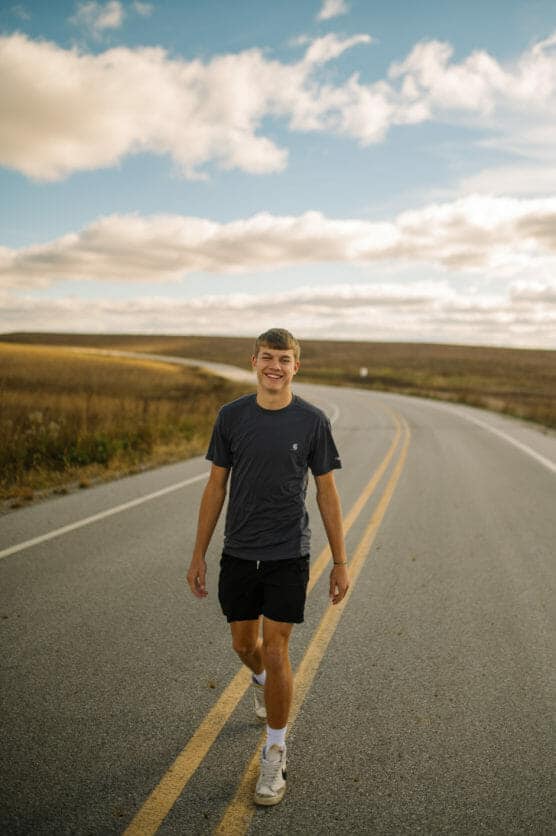 A young man in a dark t-shirt and shorts walks and smiles on an empty rural road under a partly cloudy sky, with fields stretching out on both sides. by Justin Salem Meyer