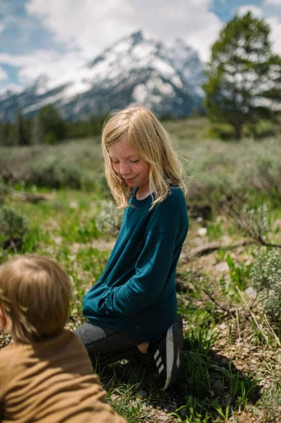 A blonde child in a blue sweater kneels outdoors on grassy ground, looking down, with mountains and pine trees in the background. Another child in a brown shirt is in the foreground, facing away. by Justin Salem Meyer