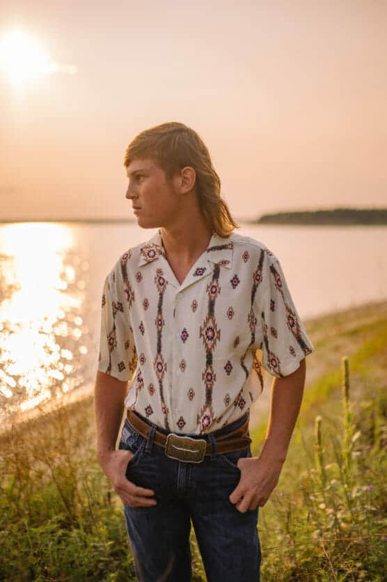 A young man with a mullet hairstyle stands outdoors near a body of water at sunset, wearing a patterned short-sleeve shirt, jeans, and a belt, gazing to the side with his hands in his pockets. by Justin Salem Meyer