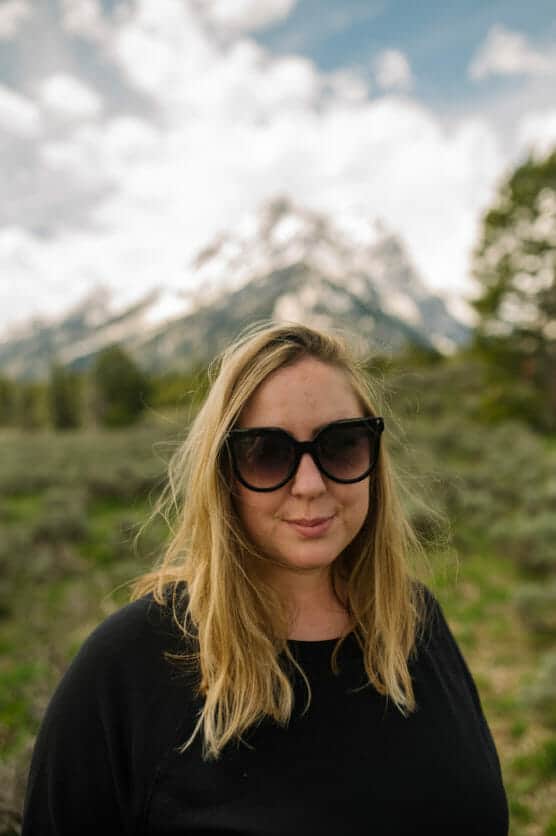 A woman with blonde hair and black sunglasses stands outdoors in front of snow-capped mountains and greenery under a partly cloudy sky. by Justin Salem Meyer