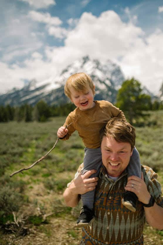 A man smiles while carrying a laughing toddler on his shoulders. The toddler holds a stick. They are outdoors in a grassy area with trees and snow-capped mountains in the background under a partly cloudy sky. by Justin Salem Meyer