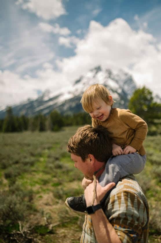 A man carries a smiling young child on his shoulders in a grassy field, with snow-capped mountains and a partly cloudy sky in the background. by Justin Salem Meyer