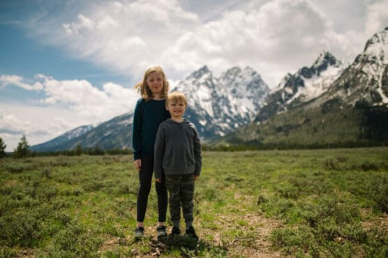 Two children stand on grassy terrain with snow-capped mountains and a cloudy sky in the background. The older child stands behind the younger, both facing the camera and smiling. by Justin Salem Meyer