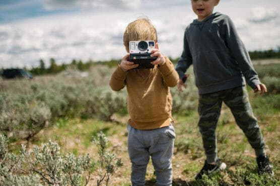 A young child holds a vintage instant camera up to their face, ready to take a photo, while another child in a gray hoodie and camouflage pants stands nearby in an open field with green shrubs. by Justin Salem Meyer
