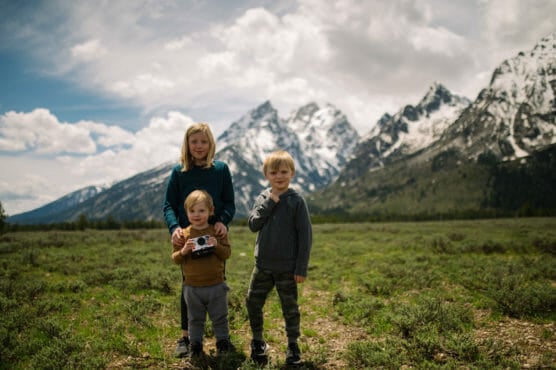 Three children stand on a grassy field with snow-capped mountains in the background. The youngest holds a camera, while the other two smile. The sky is cloudy with patches of blue. by Justin Salem Meyer