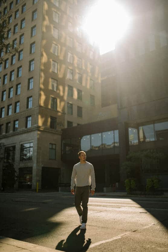 A man in casual clothing walks across a sunlit city street, casting a long shadow. Tall buildings surround him, and bright sunlight streams from above, partially obscuring the view. by Justin Salem Meyer