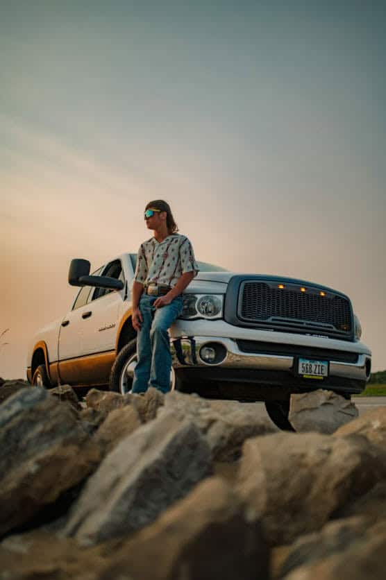 A person wearing sunglasses and a patterned shirt leans against the front of a white pickup truck, with rocks in the foreground and a soft, colorful sky in the background. by Justin Salem Meyer