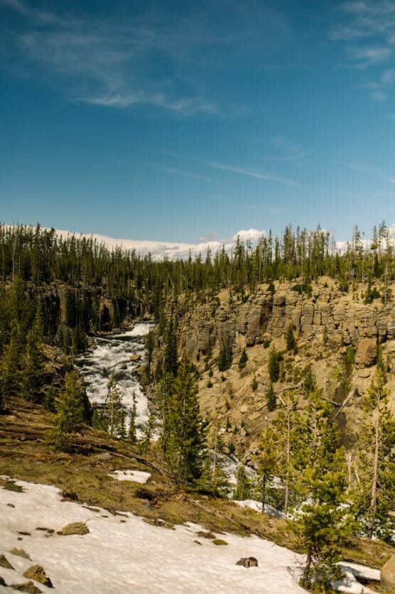 A scenic view of a rocky canyon with a river flowing through it, surrounded by pine trees and patches of snow under a partly cloudy blue sky. by Justin Salem Meyer