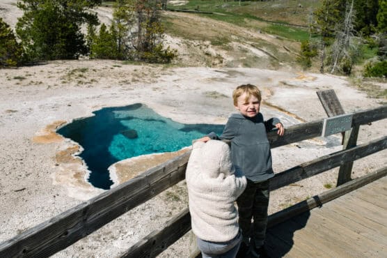 Two young children stand on a wooden boardwalk overlooking a vibrant blue hot spring surrounded by rocky ground and trees. One child faces the camera, while the other leans on the railing, facing away. by Justin Salem Meyer