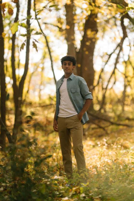 A person stands in a sunlit forest clearing, wearing a light blue shirt over a white t-shirt and khaki pants, surrounded by green and yellow foliage. by Justin Salem Meyer