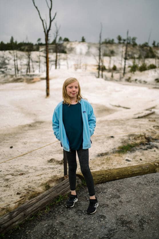 A young girl wearing a blue jacket and dark leggings stands on a path in front of a snowy or sandy landscape with bare trees in the background. by Justin Salem Meyer