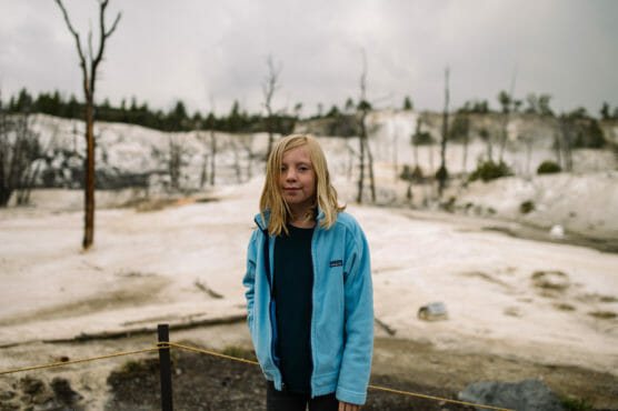 A child with long blonde hair stands outside in a pale blue jacket, with a barren, rocky landscape and leafless trees in the background under a cloudy sky. by Justin Salem Meyer