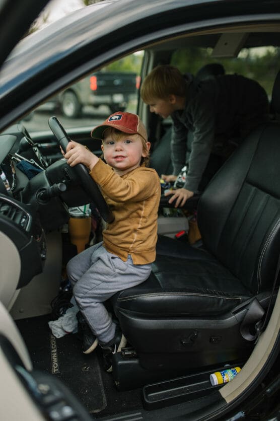 A young child wearing a brown hat and sweatshirt sits in the driver’s seat of a car, holding the steering wheel, while another child climbs in the back. The car door is open and toys are visible inside. by Justin Salem Meyer