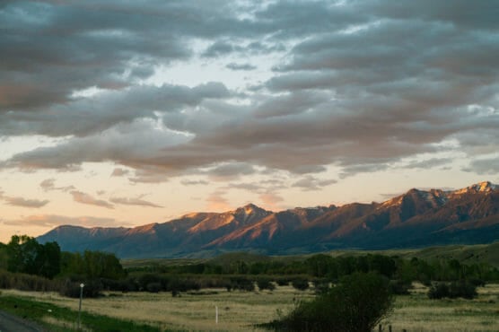 Mountain range under a cloudy sky at sunset, with sunlight illuminating the peaks. Below, there are fields, trees, and patches of green vegetation, creating a calm and serene landscape scene. by Justin Salem Meyer