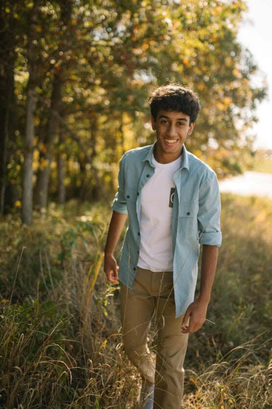 A young man with curly hair, wearing a light blue shirt over a white t-shirt and khaki pants, smiles while walking outdoors through tall grass with trees and sunlight in the background. by Justin Salem Meyer