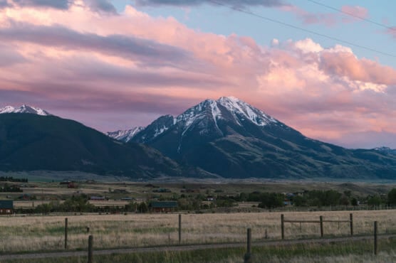 Snow-capped mountain under a colorful sunset sky with pink clouds, surrounded by green hills and a rural landscape with scattered houses, fields, and a wooden fence in the foreground. by Justin Salem Meyer