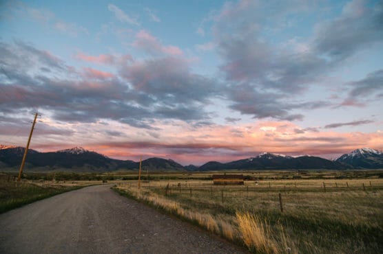 A gravel road curves through a field toward distant mountains under a sky filled with colorful sunset clouds. Snow caps the mountain peaks, and wooden fences line the grassy landscape. by Justin Salem Meyer