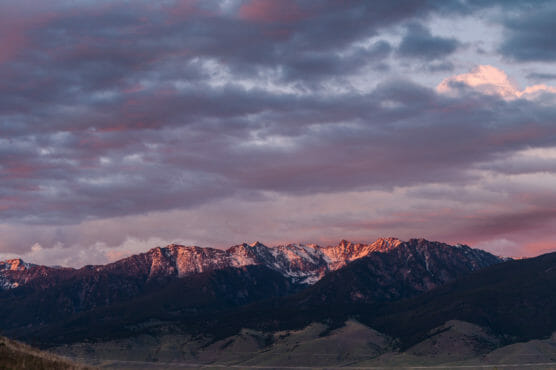 Snow-capped mountains are illuminated by the soft pink and purple hues of a sunset, with dramatic clouds filling the sky above and gentle slopes in the foreground. by Justin Salem Meyer