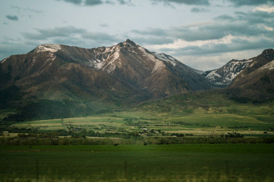 A scenic landscape featuring tall mountains with patches of snow, green rolling hills, and a wide grassy plain under a cloudy sky. Scattered trees and small buildings dot the valley below the mountains. by Justin Salem Meyer